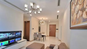 A modern living room with a flat-screen TV, a sofa, a dining table with chairs, and a chandelier. - Golden Grand Landmark (Ho Chi Minh City)