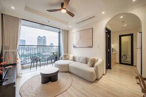 A modern living room with a sectional sofa, a round coffee table, and a view of the city through large windows. - Golden Grand Landmark (Ho Chi Minh City)