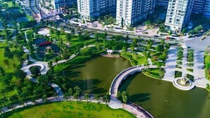 Uma área residencial com edifícios modernos e altos, um parque central com uma fonte circular e uma ponte pedonal curva.