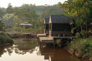 A serene riverside setting with a modern house featuring a large window and a balcony. - The Village Resort in the Painting, Bao Loc (Bao Lam District)