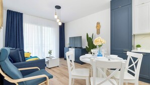 Uma sala de estar moderna com um sofá azul, cadeiras brancas, uma mesa de jantar posta para quatro pessoas e uma área de cozinha com armários azuis e bancada branca.