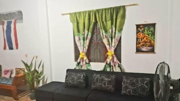 Een woonkamer met een zwarte bank, een ventilator en een groen gordijn met bloemenmotieven.