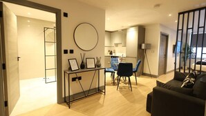 Una sala moderna con un sofá negro, un comedor con sillas azules y una cocina al fondo.