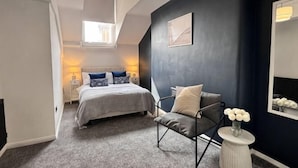 A bedroom with a bed, a chair, a small table with flowers, a mirror, and a window.