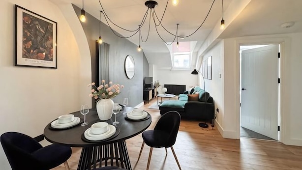 A modern dining area with a round table, black chairs, and a vase of flowers. There is a sofa and a door in the background.