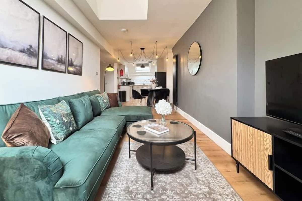 A modern living room with a green sofa, a round coffee table, a TV, and a kitchen in the background.