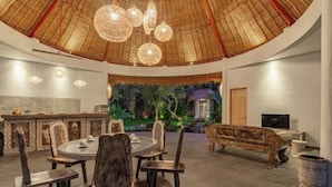 Una habitación amplia con una mesa de comedor redonda, sillas de madera, un sofá y un gran ventanal con vistas a un jardín exuberante.