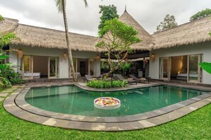 A pool with a circular float, surrounded by a stone deck, with a thatched-roof building and a covered seating area in the background. - Brand New luxury 3BR Villa Ethnic Ubud #2 (Bali)