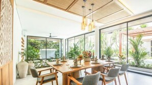 A spacious dining area with a long wooden table, multiple chairs, and large windows offering a view of greenery outside. - Swastamita (Bali)