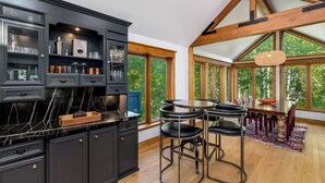 A modern kitchen with dark cabinetry, a marble countertop, and a dining area with a wooden table and bar stools. - Evergreen Escape - Home for 10 (Vail)