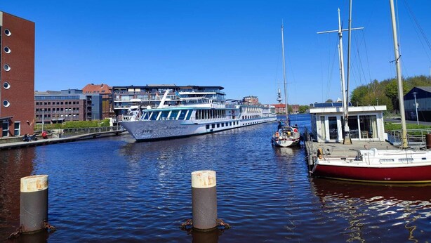 Une rivière où est amarré un grand bateau à un quai, un plus petit bateau se trouvant à proximité, et un bâtiment à la façade en briques rouges en arrière-plan.