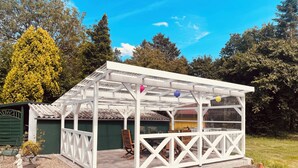 Un espace extérieur aménagé avec des sièges, abrité par un toit blanc et une structure en treillis, et entouré de verdure et d’arbres.