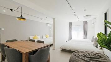 A modern hotel room with a wooden dining table, grey chairs, a large bed, and a bedside plant.