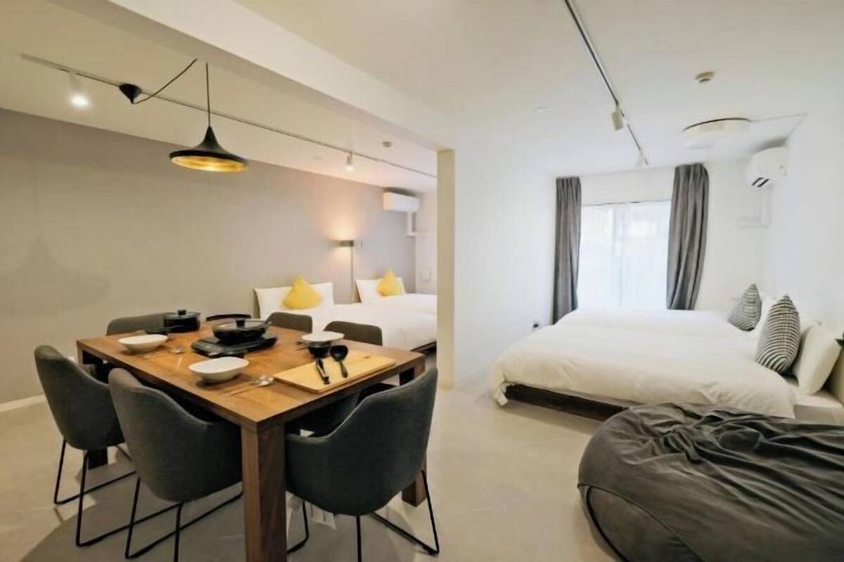 A modern hotel room with a wooden dining table, gray chairs, a bed with white bedding, and a gray bean bag.