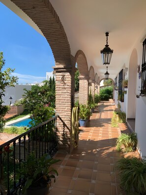 A covered outdoor corridor with brick arches, potted plants, and a pool.