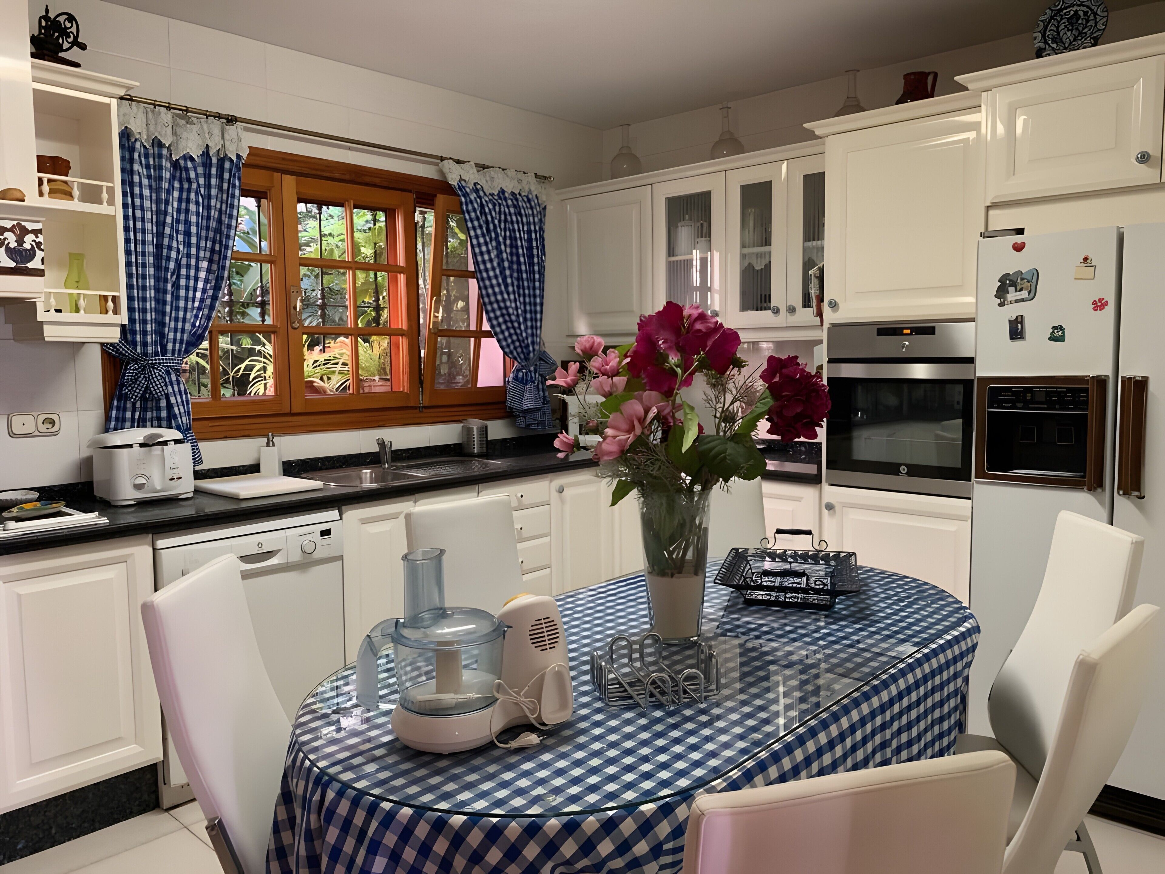 Una cocina con armarios blancos, una ventana con cortinas, una mesa de comedor con mantel a cuadros y un florero con flores.