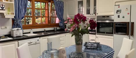 Una cocina con armarios blancos, una ventana con cortinas, una mesa de comedor con mantel a cuadros y un florero con flores.