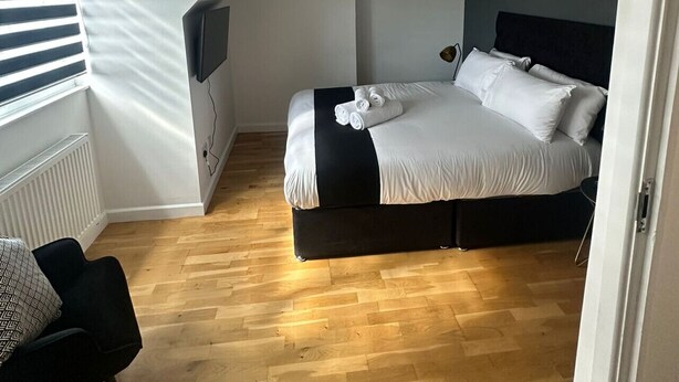 Camera da letto con soffitto a falde, letto con lenzuola bianche, poggiatesta nero, una sedia nera e lampadario.