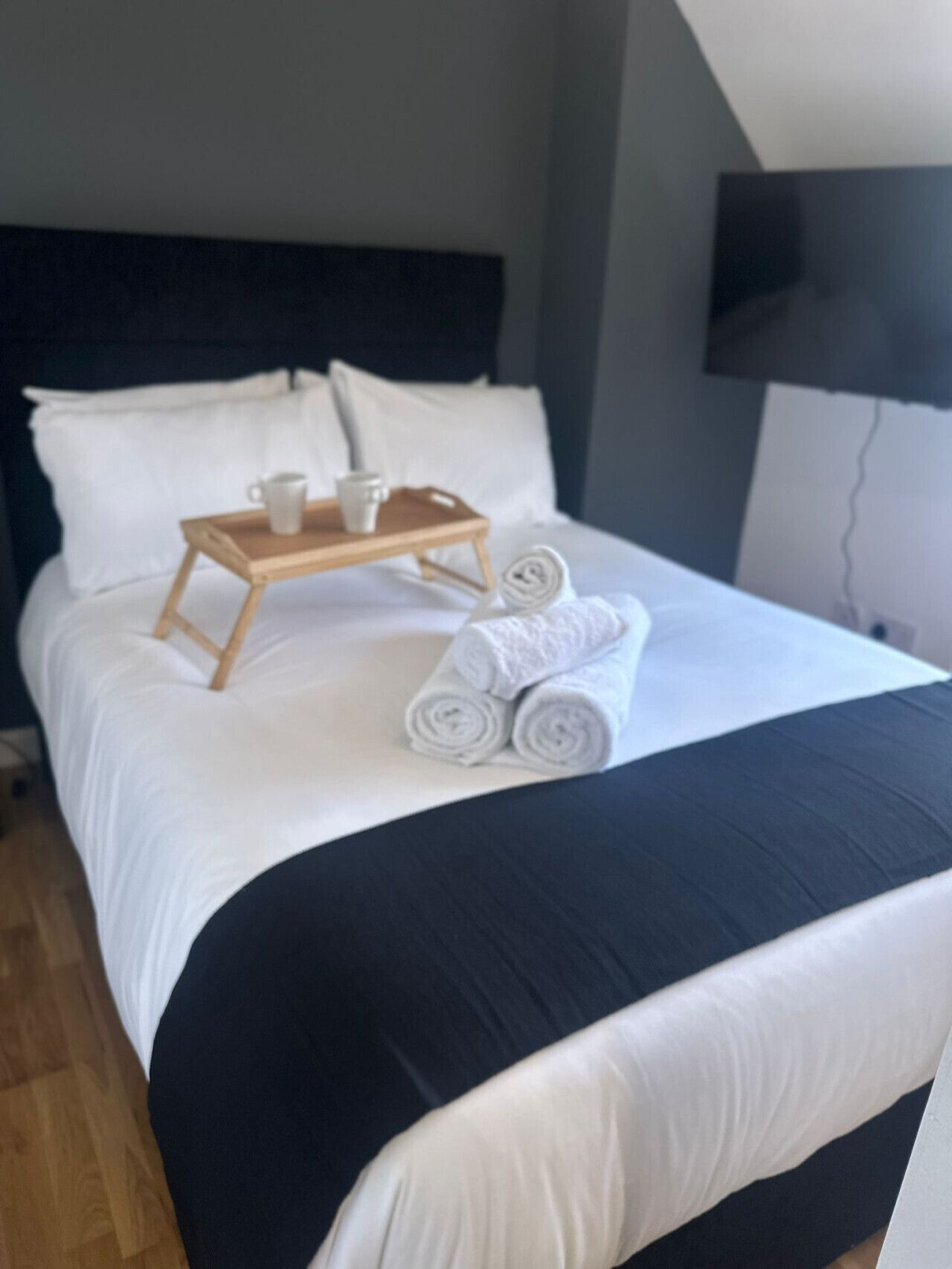 Una cama bien hecha con sábanas blancas, una bandeja de madera con dos tazas y toallas enrolladas.