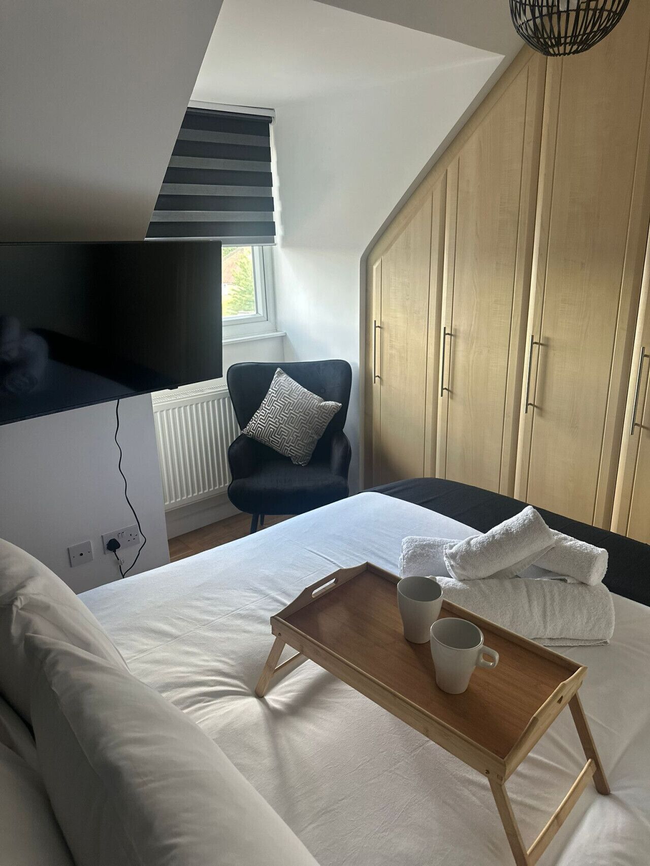 A bedroom with a bed, a wooden bedside table with a tray and a cup, a black chair with a patterned pillow, a wardrobe, and a window with blinds.