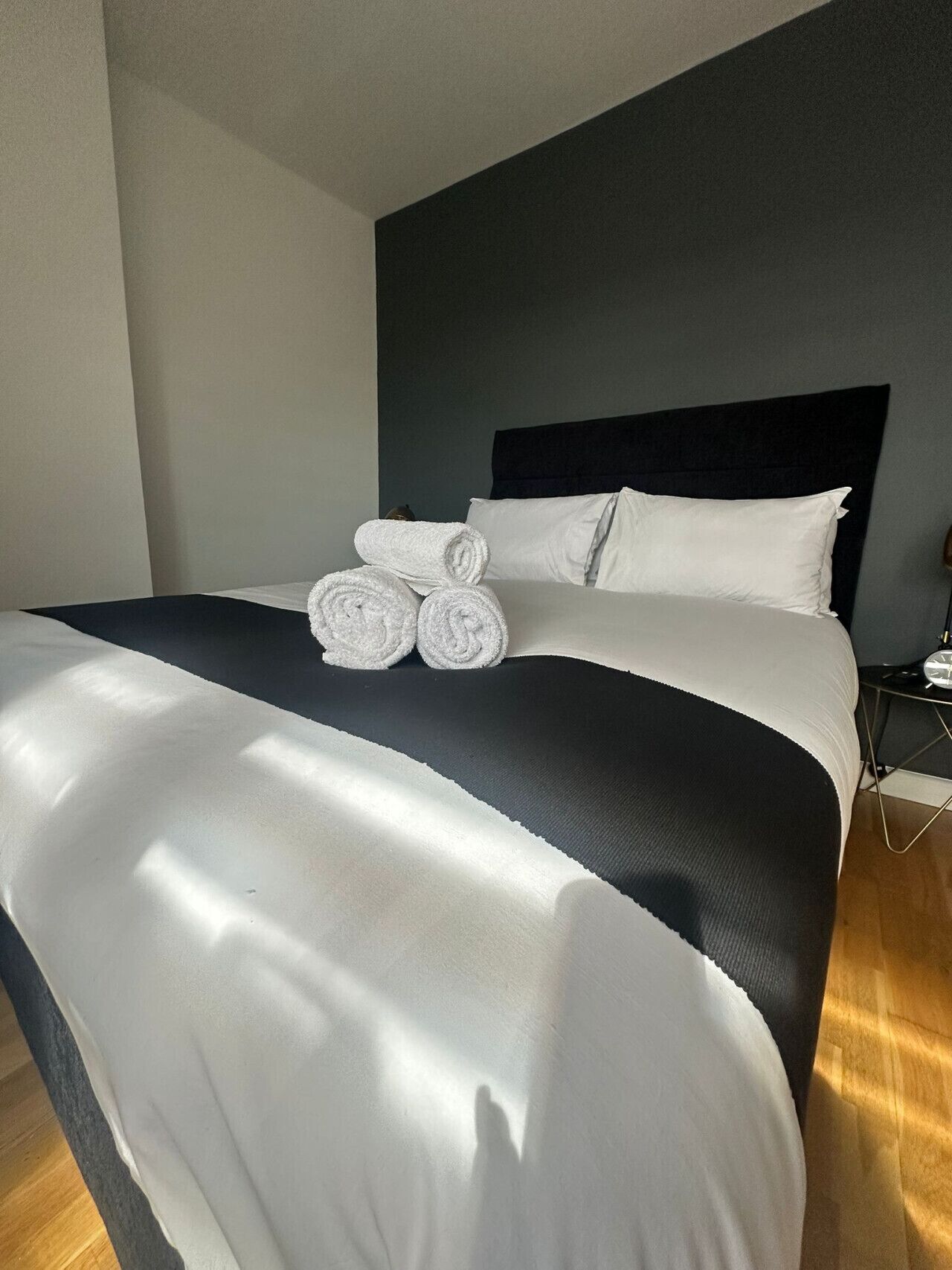 A neatly made bed with white linens and rolled towels, a dark headboard, and a bedside table with a clock.