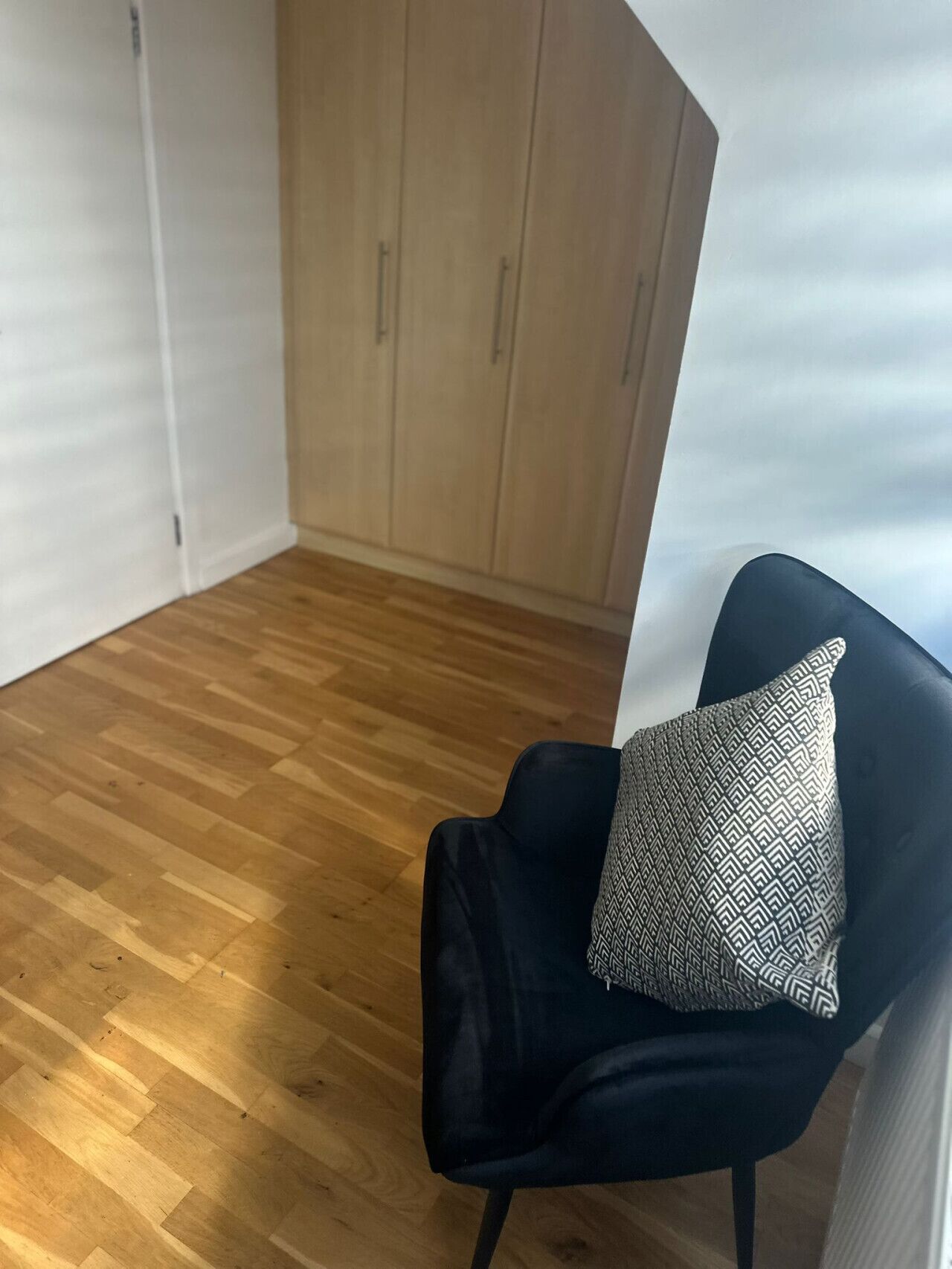 Una habitación con piso de madera, una silla negra con un cojín estampado y un armario blanco.
