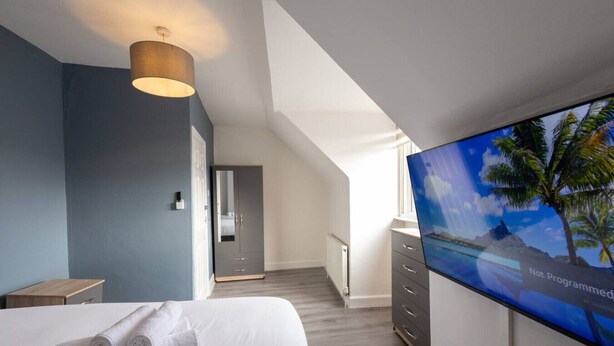 Een moderne hotelkamer met een bed, een televisie waarop een tropisch strand te zien is, een garderobe en een gang naar een andere kamer.