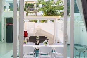 Une chambre d’hôtel avec un balcon, un coin repas avec des chaises blanches et une table, et une vue sur les bâtiments environnants et la verdure.