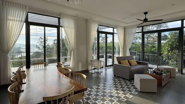 A spacious room with a large dining table, comfortable seating, and large windows offering a view of greenery. - MyGarden Nam Phuong Lakeview (Bao Loc)