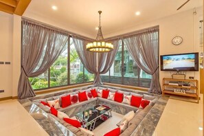 Una sala de estar con un sofá seccional, una mesa de centro, un televisor y ventanas amplias con cortinas.
