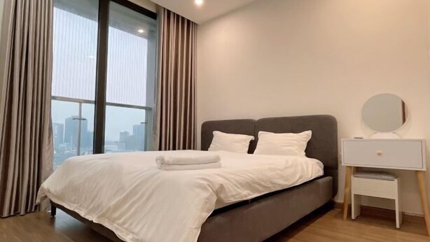 A modern hotel room with a large bed, a nightstand, and a view of the cityscape. - Warm 4-Bedroom Apartment At Vinhomes Skylake (Hanoi)