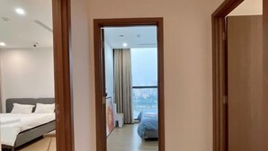 A hotel room with a bed, a small table, and a view of the city through large windows. - Warm 4-Bedroom Apartment At Vinhomes Skylake (Hanoi)