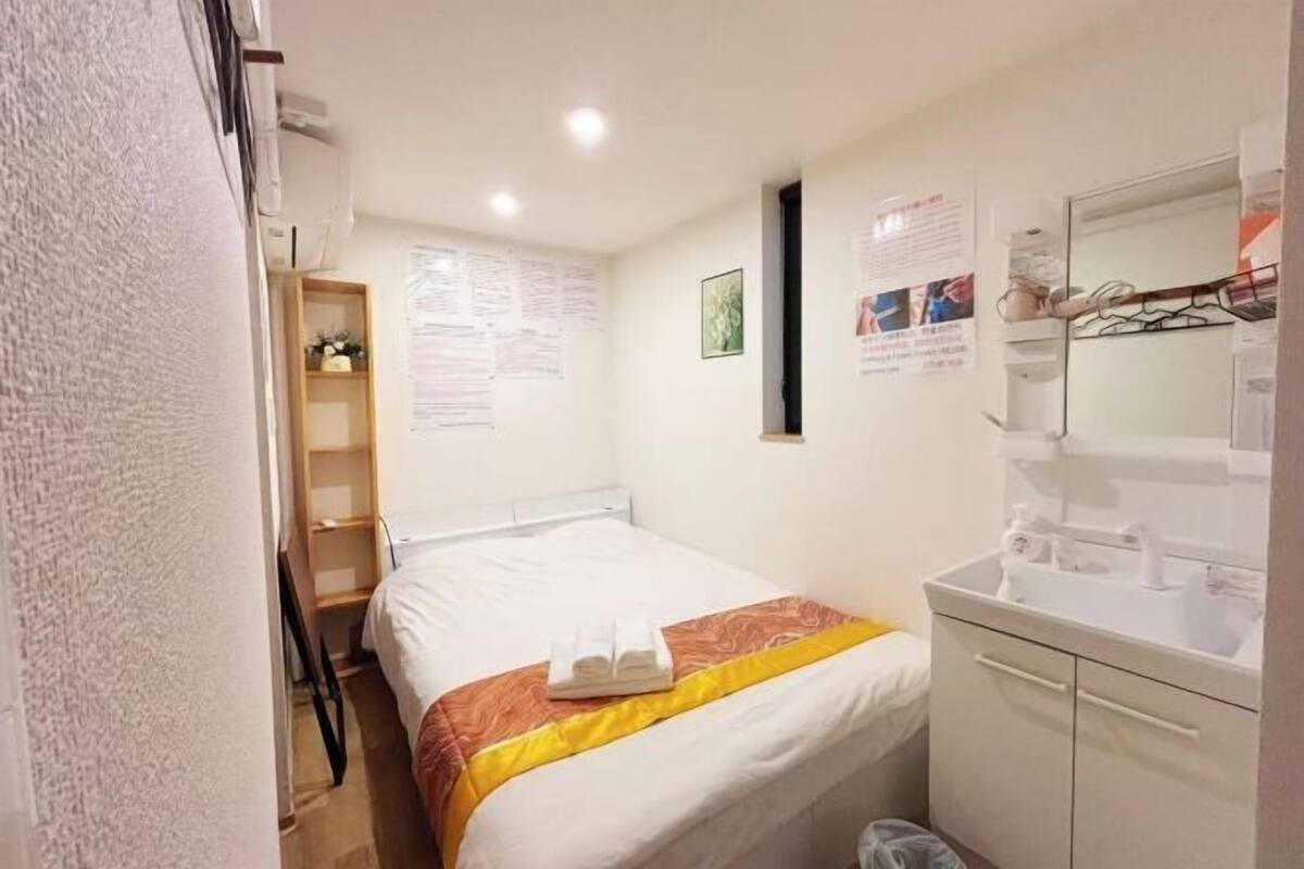 Una habitación pequeña y limpia con cama, mesita de noche, espejo y un estante con artículos de aseo.
