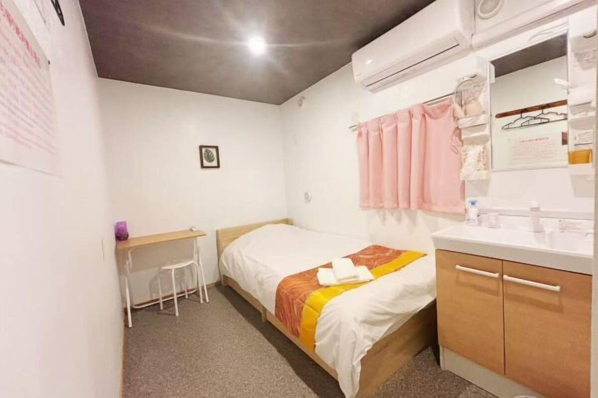 Una habitación de hotel pequeña con una cama, un escritorio, una silla, un baño con espejo y un aire acondicionado instalado en la pared.