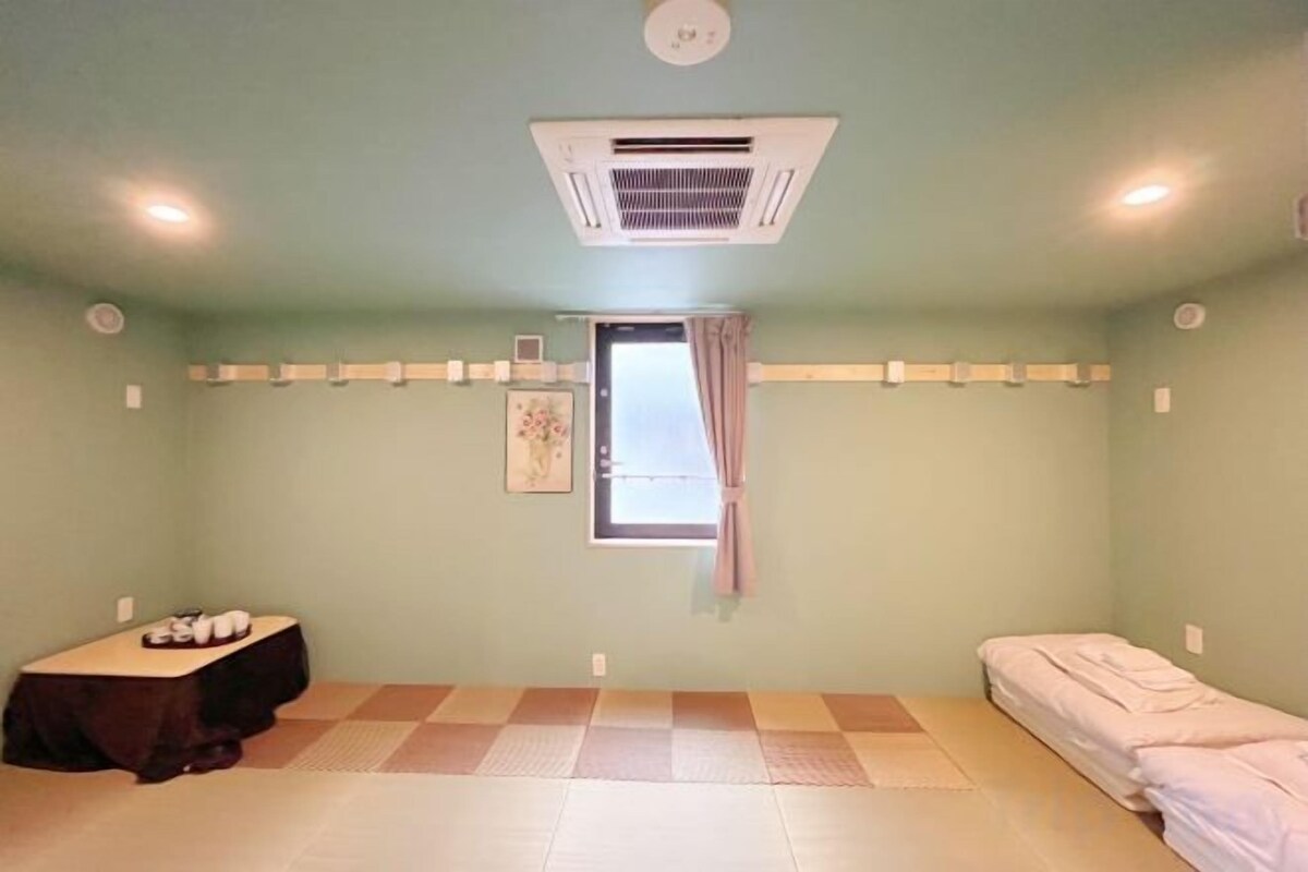 A room with a bed, a small table, a window with curtains, and a ceiling-mounted air conditioner.