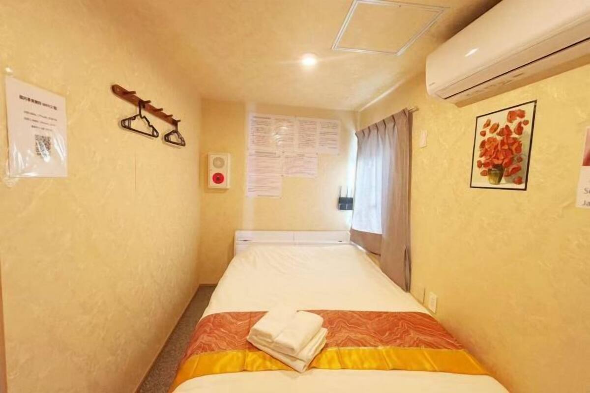 Una habitación de hotel con cama, aire acondicionado de pared, perchero y un cuadro enmarcado.