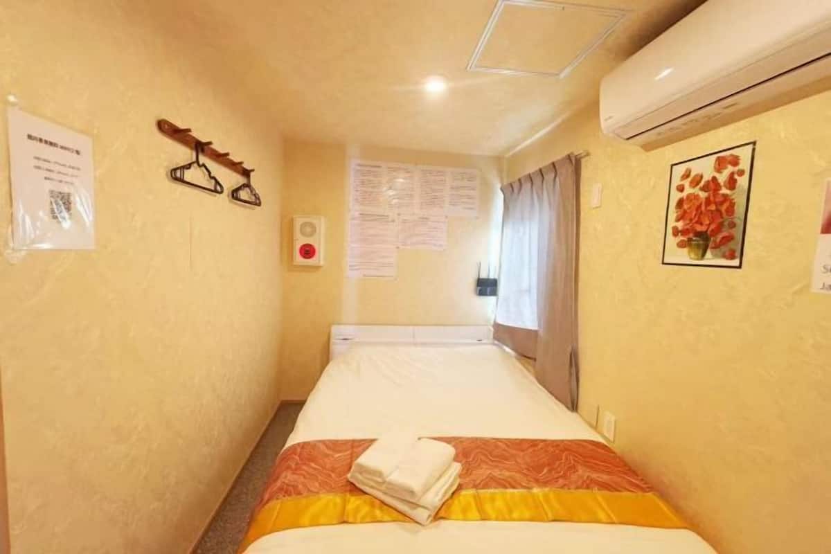 A hotel room with a bed, a wall-mounted air conditioner, a coat rack, and a framed picture.