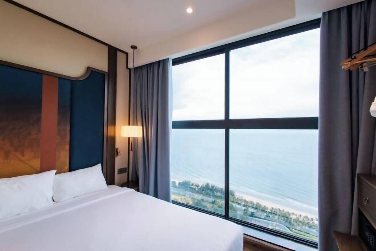 Habitación de hotel con una cama grande, una mesita de noche, una lámpara y vista al mar a través de un ventanal amplio.