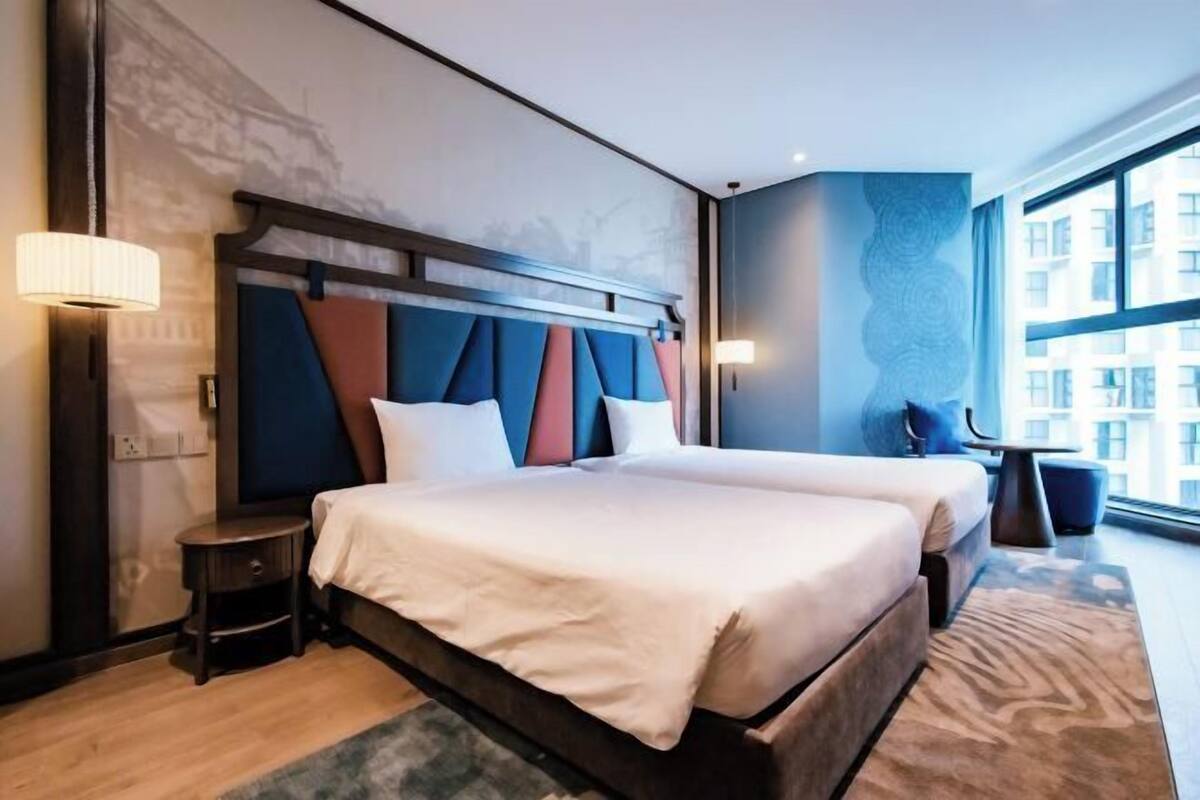 Una habitación de hotel moderna con una cama grande, dos mesitas de noche y vistas a la ciudad a través de amplios ventanales.