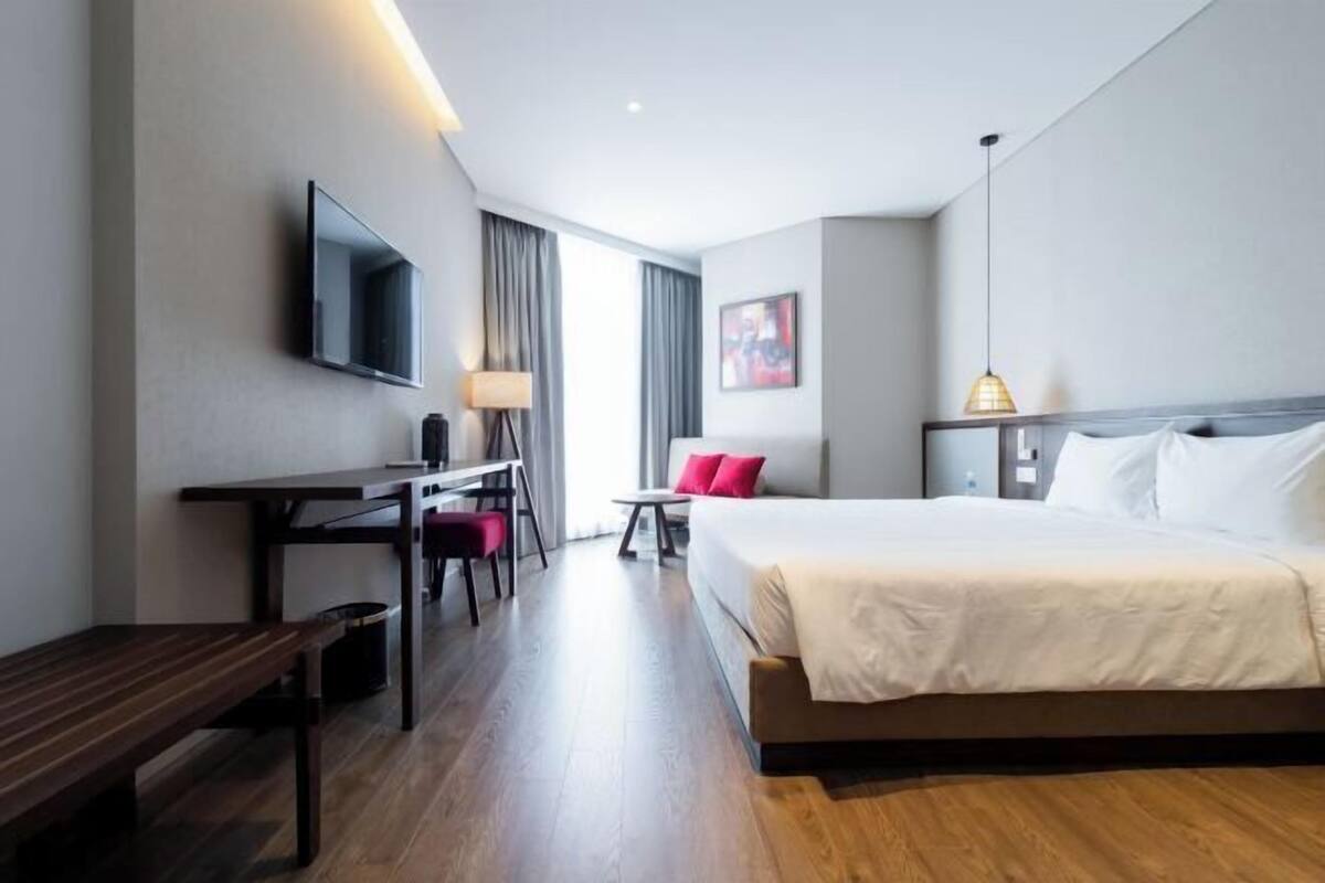Habitación de hotel con cama, escritorio, silla, televisor, lámpara y una obra de arte en la pared.