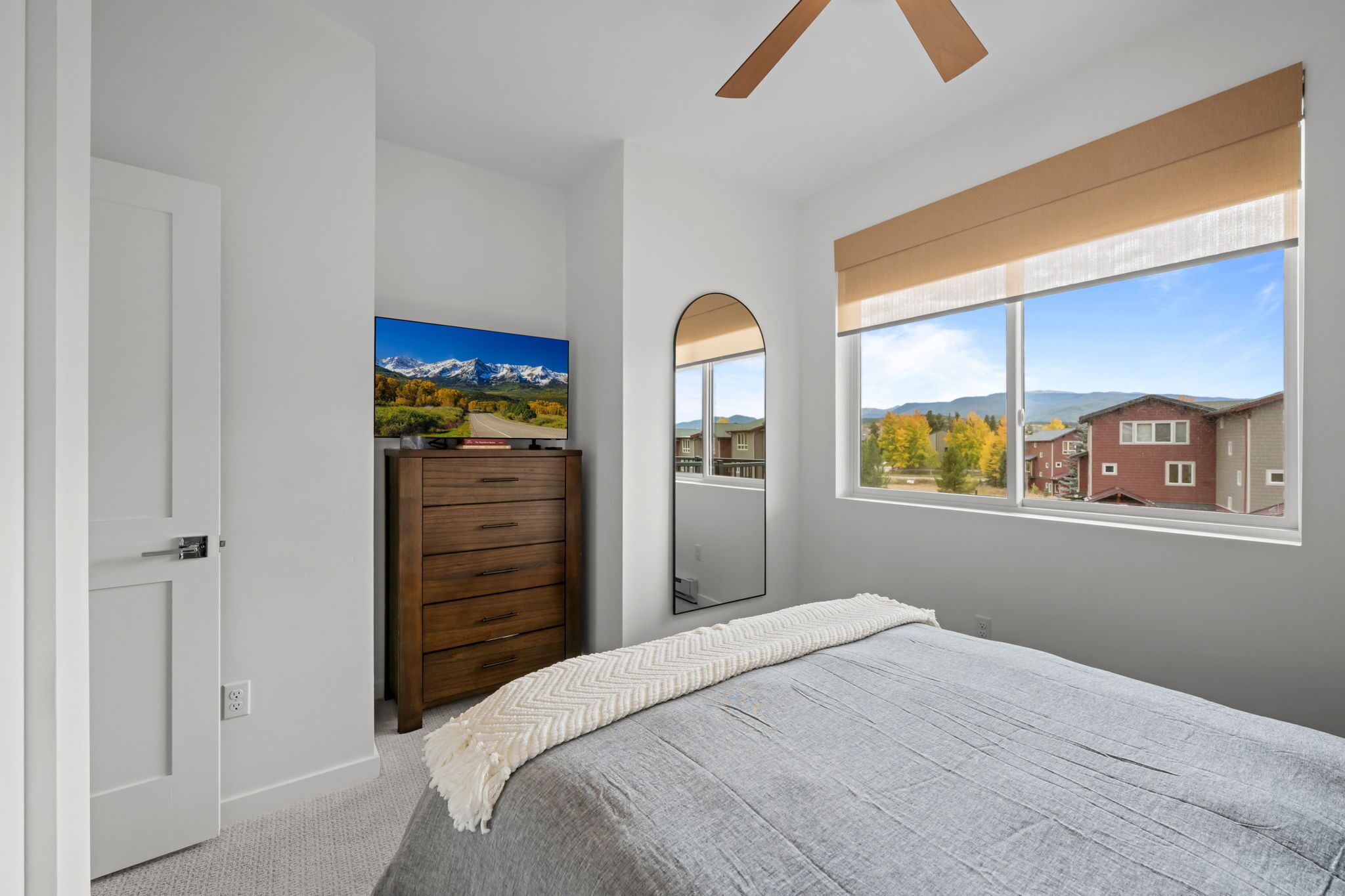 A bedroom with a bed, a dresser, a painting, a mirror, and a window with a view of a house and mountains.