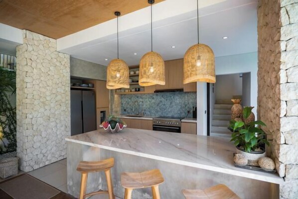 A modern kitchen with a marble island, wooden stools, and hanging wicker lights.