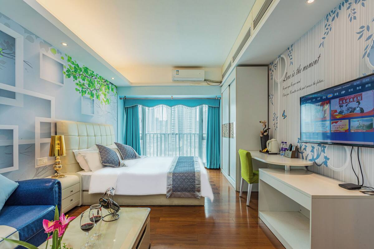 Ein modernes Hotelzimmer mit einem großen Bett, einem Schreibtisch mit Computer, einem Fernseher und Blick auf die Stadt durch große Fenster.