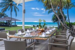 A dining area with tables set for a meal, overlooking a beach with palm trees and a clear sky.