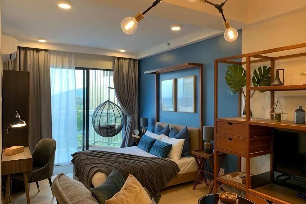 A bedroom with a bed, desk, chair, hanging chair, and a large window.