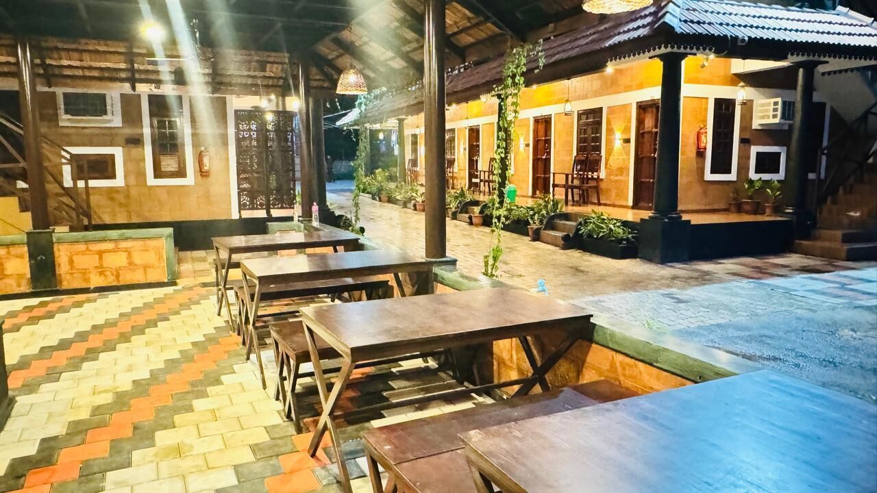 Un espace extérieur abrité, avec des tables et des chaises en bois, un sol carrelé et, en arrière-plan, un bâtiment aux murs en bois et au toit recouvert de tuiles.