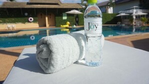 A poolside setting with a towel and a bottle of Joy water, a pool, and a building with a thatched roof in the background. - Baan PromTel , Luxury Villa close to Nimman, Resort Pool, 4BR (Chiang Mai)