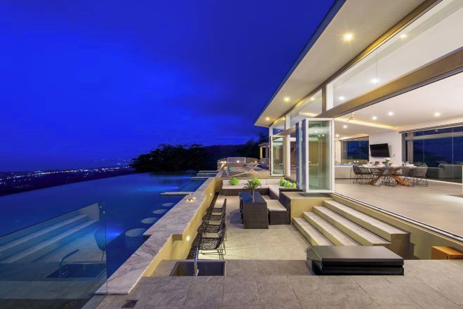 A modern house with a swimming pool, outdoor seating, and a view of the city at dusk.