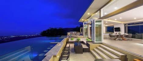 A modern house with a swimming pool, outdoor seating, and a view of the city at dusk.