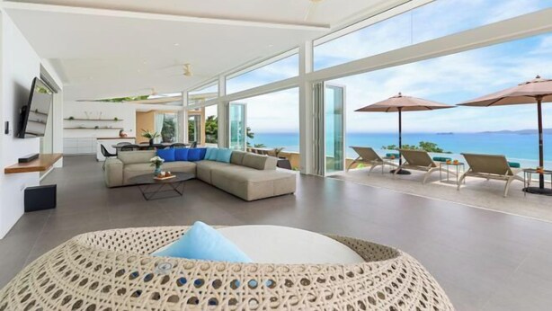 A spacious living area with a large sofa, a coffee table, and a view of the ocean. There are several lounge chairs with umbrellas outside. - Villa Cabo: Hilltop Retreat with Panoramic Ocean Views (Koh Samui)